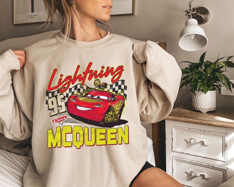 Puede incluir: Sudadera beige con un gr&aacute;fico de Rayo McQueen de la pel&iacute;cula Cars. El dise&ntilde;o incluye el texto "Lightning McQueen 95 Cars" en rojo y amarillo. La sudadera tiene mangas largas y cuello redondo.