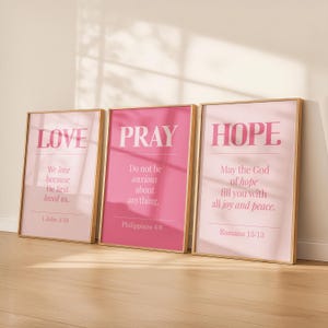 May include: Three rectangular wall art prints in light wooden frames, each featuring pink backgrounds and white text. The prints display the words LOVE, PRAY, and HOPE, accompanied by Bible verses 1 John 4:19, Philippians 4:6, and Romans 15:13.