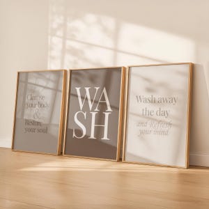 Peut inclure: Trois affiches murales verticales encadrées avec des cadres en bois clair. L'affiche de gauche est taupe avec le texte blanc Cleanse your body & Restore your soul. Celle du centre est marron foncé avec le texte blanc WASH. Celle de droite est blanche avec le texte taupe Wash away the day and Refresh your mind.