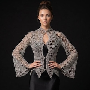 May include: A silver chainmail top with long flared sleeves, a high collar, and a pointed hemline, worn over a floor-length black satin slip dress. The metallic top features a fine interlocking ring pattern and a front opening secured with two clasps.