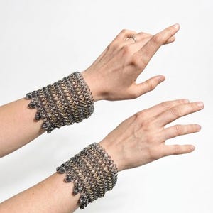 May include: Two chainmail wrist cuffs, each made of small, interlocking metal rings. The cuffs have a textured appearance and a silver and gold colour scheme. The cuffs are worn on the wrists, with the hands in the background.