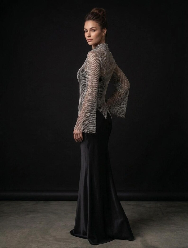 May include: A long-sleeved top made of silver-toned metallic chainmail with a high neckline and flared cuffs, paired with a floor-length black skirt. The chainmail garment features a repeating interlocking ring pattern and a pointed hemline.