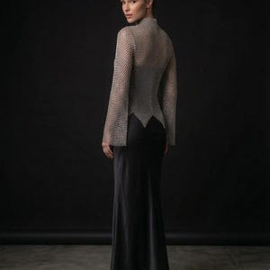 May include: A long-sleeved silver chainmail top with a high neckline and a pointed hemline, worn over a floor-length black satin slip dress. The metallic mesh garment features wide sleeves and a fitted silhouette that contrasts with the smooth fabric.