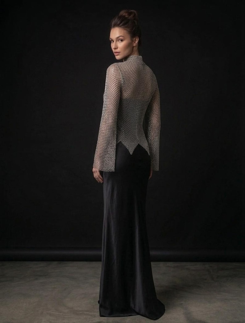 May include: A long-sleeved silver chainmail top with a high neckline and a pointed hemline, worn over a floor-length black satin slip dress. The metallic mesh garment features wide sleeves and a fitted silhouette that contrasts with the smooth fabric.