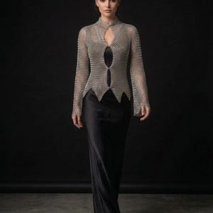 May include: A long-sleeved silver chainmail top featuring a high collar and a front cutout design, layered over a floor-length black satin dress. The metallic mesh garment has a pointed hemline and long sleeves, providing a structured contrast to the smooth, flowing black fabric of the full-length dress underneath.