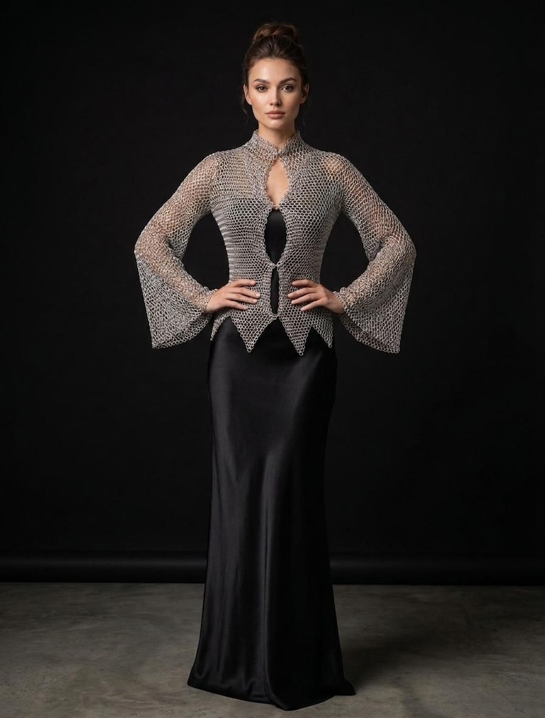 May include: A silver chainmail top with long flared sleeves, a high collar, and a pointed hemline, worn over a floor-length black satin slip dress. The metallic top features a fine interlocking ring pattern and a front opening secured with two clasps.