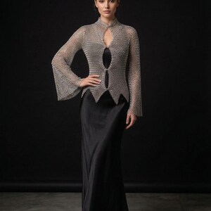 May include: A long-sleeved silver chainmail top with a high collar and a pointed hemline, worn over a floor-length black satin slip dress. The metallic top features a front opening with two circular closures, while the dress has a smooth finish.
