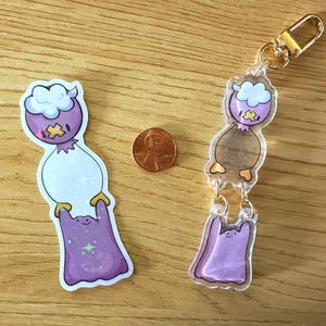 May include: A sticker and a keychain featuring a cartoon character with a purple head, white body, and purple cape. The keychain has a gold clasp. A penny is next to the items for size comparison.