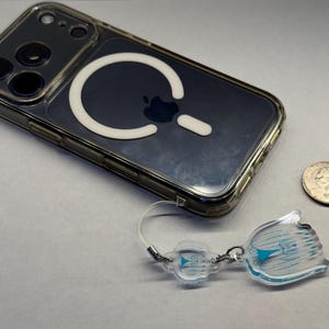 May include: A clear phone case with a white MagSafe ring and a clear phone charm with a blue design. A quarter is next to the phone. The phone case is on a white surface.