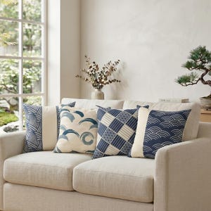 May include: Four square throw pillows arranged on a light beige sofa. The pillows feature various blue and white patterns, including geometric grids, wave motifs, and abstract circular designs, all set against a neutral, light-colored background.