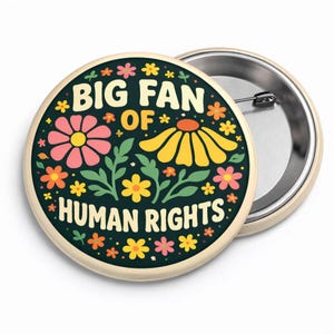 May include: A round button with the text "BIG FAN OF HUMAN RIGHTS" in white and yellow, surrounded by colorful flowers. The button has a dark green background and a metal pin back.