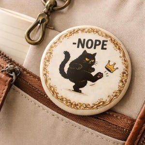 May include: A circular badge featuring a black cat kicking a gold crown, with the word "-NOPE" above. The badge has a gold floral border. The background is a neutral colour.