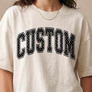 May include: An off-white short-sleeve t-shirt featuring the word CUSTOM printed in a large, arched, black collegiate-style font. The letters are filled with a white polka dot pattern and outlined in black, creating a bold, high-contrast graphic design.