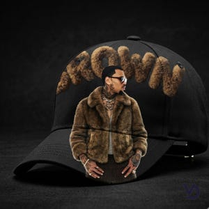Puede incluir: Gorra de b&eacute;isbol negra con la palabra "BROWN" en letras de piel sint&eacute;tica marr&oacute;n. La gorra presenta una imagen de una persona que lleva un abrigo de piel marr&oacute;n y gafas de sol. La gorra tiene una visera curva.