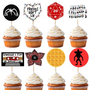May include: Cupcakes with white frosting and various themed toppers. Toppers include a black silhouette, a heart with the text "FRIENDS DON'T LIE", a red D20 die, and the alphabet with string lights. Other toppers include a cassette tape, a Demogorgon flower, a waffle, and a Demogorgon silhouette.