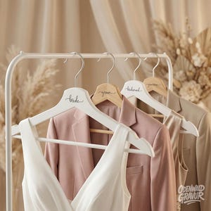Könnte beinhalten: Weiße und hölzerne Kleiderbügel mit den Wörtern "bride", "groom" und "bridesmaid" in Schreibschrift. Ein weißes Kleid und rosa und beige Blazer hängen an den Bügeln. Die Bügel befinden sich auf einem weißen Gestell, mit neutralem Hintergrund.