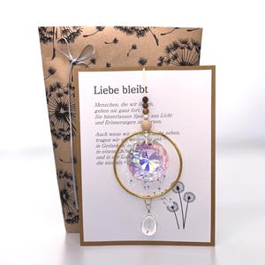 Könnte beinhalten: Ein dekorativer Hängeschmuck mit einem goldfarbenen Metallring, einer facettierten, irisierenden Kristallkugel und einem kleinen, klaren ovalen Anhänger mit einer Pusteblumensamen-Einlage vor einer Karte mit dem Text Liebe bleibt.
