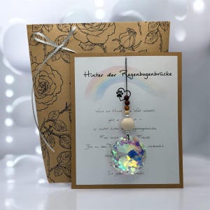 Könnte beinhalten: Dekorativer Kristallprismen-Anhänger mit Regenbogeneffekt, hängend an einer Karte mit deutschem Text und einem Hundepfotenabdruck. Die Karte befindet sich vor einer braunen Papiertüte mit Rosen.