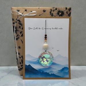Könnte beinhalten: Eine Grußkarte mit einem Berglandschaftsdesign und dem deutschen Text "Das Licht der Erinnerung leuchtet weiter." Eine dekorative Kristallkugel hängt an der Karte. Die Karte befindet sich neben einem verpackten Geschenk.