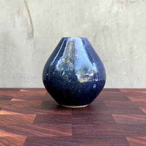 May include: A small, dark blue ceramic vase with a glossy finish. The vase has a rounded body and a narrow neck. The dark blue color has a speckled appearance. The vase is sitting on a dark wood surface.