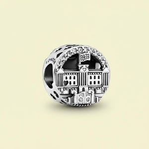 May include: A silver charm with a London theme. The charm features a detailed depiction of a building with a flag, surrounded by sparkling stones. The word "LONDON" is engraved on the side.