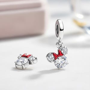 Peut inclure: Deux breloques en argent avec un design de Minnie Mouse. Chaque breloque a une grosse pierre de zircone cubique transparente pour le visage, deux petites pierres pour les oreilles et un n&oelig;ud rouge. Une breloque est suspendue, l'autre est &agrave; plat.
