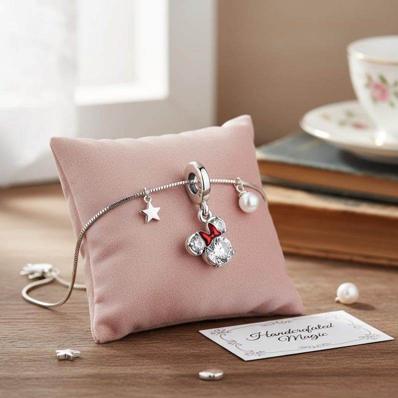 Peut inclure: Un bracelet &agrave; breloques argent&eacute; avec un charm Minnie Mouse, un charm &eacute;toile et un charm perle. Le charm Minnie Mouse a un n&oelig;ud rouge et des d&eacute;tails scintillants. Le bracelet est pr&eacute;sent&eacute; sur un coussin rose avec une carte "Handcrafted Magic".