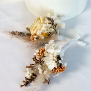 K&ouml;nnte beinhalten: Zwei kleine Ansteckblumen aus Trockenblumen. Jede Ansteckblume hat eine Mischung aus wei&szlig;en, cremefarbenen und braunen Trockenblumen mit Akzenten aus beigem Federgras. Die Stiele sind mit wei&szlig;em Band umwickelt.