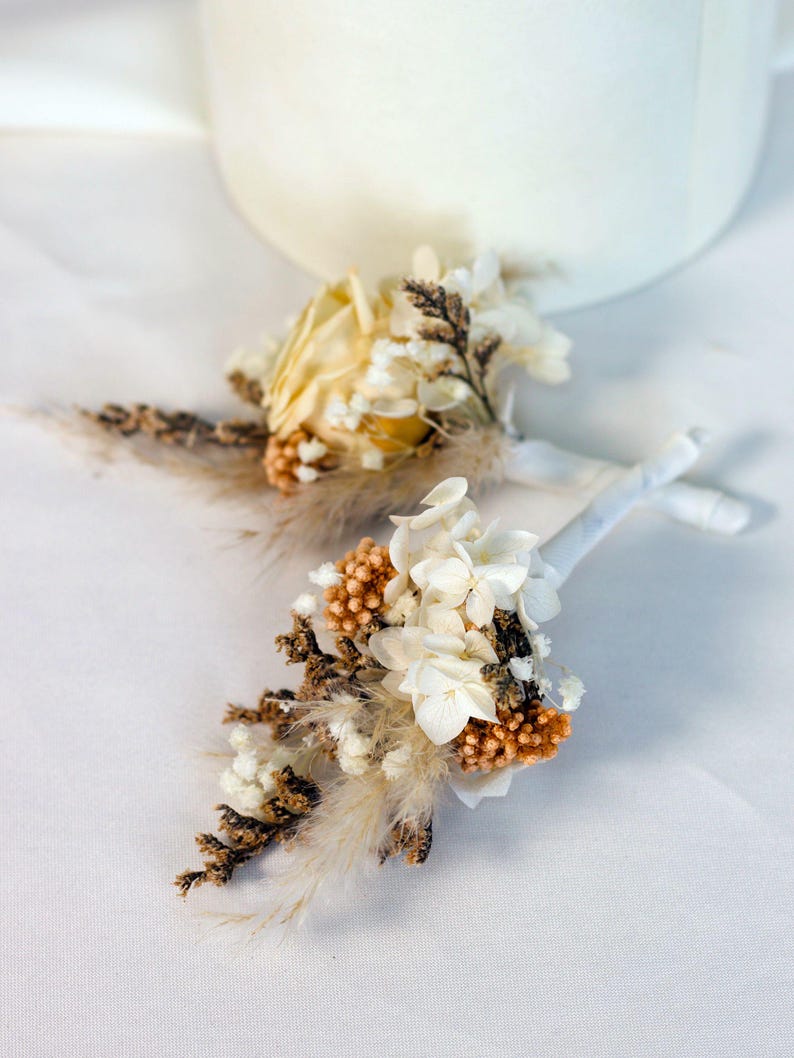 K&ouml;nnte beinhalten: Zwei kleine Ansteckblumen aus Trockenblumen. Jede Ansteckblume hat eine Mischung aus wei&szlig;en, cremefarbenen und braunen Trockenblumen mit Akzenten aus beigem Federgras. Die Stiele sind mit wei&szlig;em Band umwickelt.
