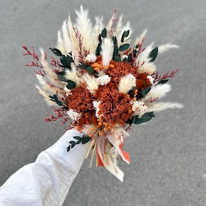 May include: A bouquet of dried flowers featuring deep rust-coloured carnations, fluffy white pampas grass, dark green eucalyptus leaves, and small sprigs of red filler flowers. The arrangement is tied with long, flowing orange and white ribbons.