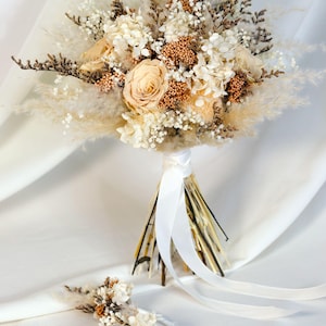 K&ouml;nnte beinhalten: Ein Brautstrau&szlig; und ein Anstecker mit Trockenblumen in Creme-, Beige- und Braunt&ouml;nen. Der Strau&szlig; enth&auml;lt Rosen, Pampasgras und strukturierte Elemente, gebunden mit einem wei&szlig;en Band. Der Anstecker spiegelt das Design des Strau&szlig;es wider.