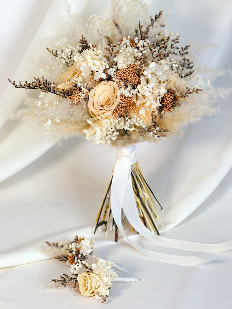 K&ouml;nnte beinhalten: Ein Brautstrau&szlig; und ein Anstecker mit Trockenblumen in Creme-, Beige- und Braunt&ouml;nen. Der Strau&szlig; enth&auml;lt Rosen, Pampasgras und strukturierte Elemente, gebunden mit einem wei&szlig;en Band. Der Anstecker spiegelt das Design des Strau&szlig;es wider.