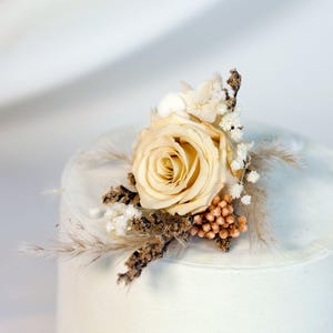 K&ouml;nnte beinhalten: Eine cremefarbene Rosen-Boutonniere mit getrockneten floralen Akzenten. Das Arrangement umfasst kleine wei&szlig;e Bl&uuml;ten, hellbraunes Gras und eine Ansammlung kleiner, runder, orange-brauner Knospen. F&uuml;r eine Hochzeit oder einen besonderen Anlass.