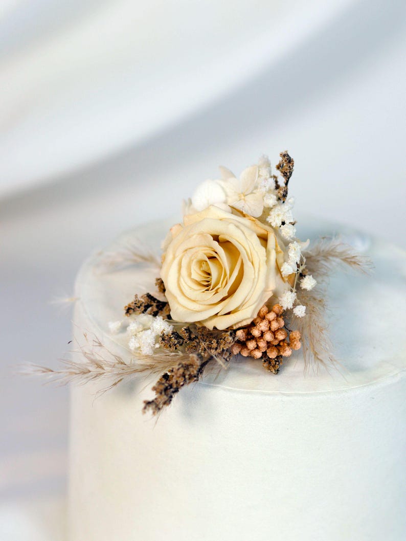K&ouml;nnte beinhalten: Eine cremefarbene Rosen-Boutonniere mit getrockneten floralen Akzenten. Das Arrangement umfasst kleine wei&szlig;e Bl&uuml;ten, hellbraunes Gras und eine Ansammlung kleiner, runder, orange-brauner Knospen. F&uuml;r eine Hochzeit oder einen besonderen Anlass.