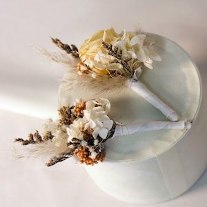 K&ouml;nnte beinhalten: Zwei kleine Trockenblumenstr&auml;u&szlig;e mit wei&szlig; bandagierten Stielen. Die Str&auml;u&szlig;e weisen cremefarbene Rosen, wei&szlig;e Hortensien und Akzente aus braunen und beigen getrockneten Gr&auml;sern und Beeren auf. Die Str&auml;u&szlig;e sind auf einer wei&szlig;en, abgerundeten Oberfl&auml;che angeordnet.