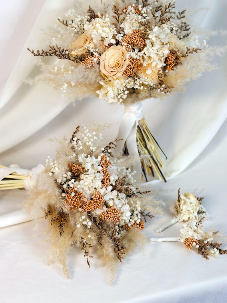 K&ouml;nnte beinhalten: Zwei Brautstr&auml;u&szlig;e und ein Knopfloch mit Trockenblumen. Die Str&auml;u&szlig;e haben eine Mischung aus beigem Pampasgras, wei&szlig;en und beigen Blumen und braunen Akzenten. Die Knopflochblume hat &auml;hnliche Elemente.