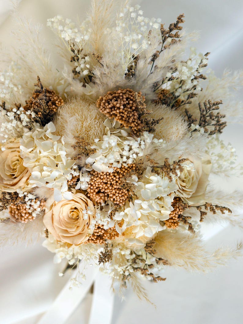 K&ouml;nnte beinhalten: Nahaufnahme eines Trockenblumenstrau&szlig;es mit cremefarbenen Rosen, Pampasgras und kleinen wei&szlig;en Bl&uuml;ten. Der Strau&szlig; hat eine nat&uuml;rliche Farbpalette mit braunen und beigen Akzenten.