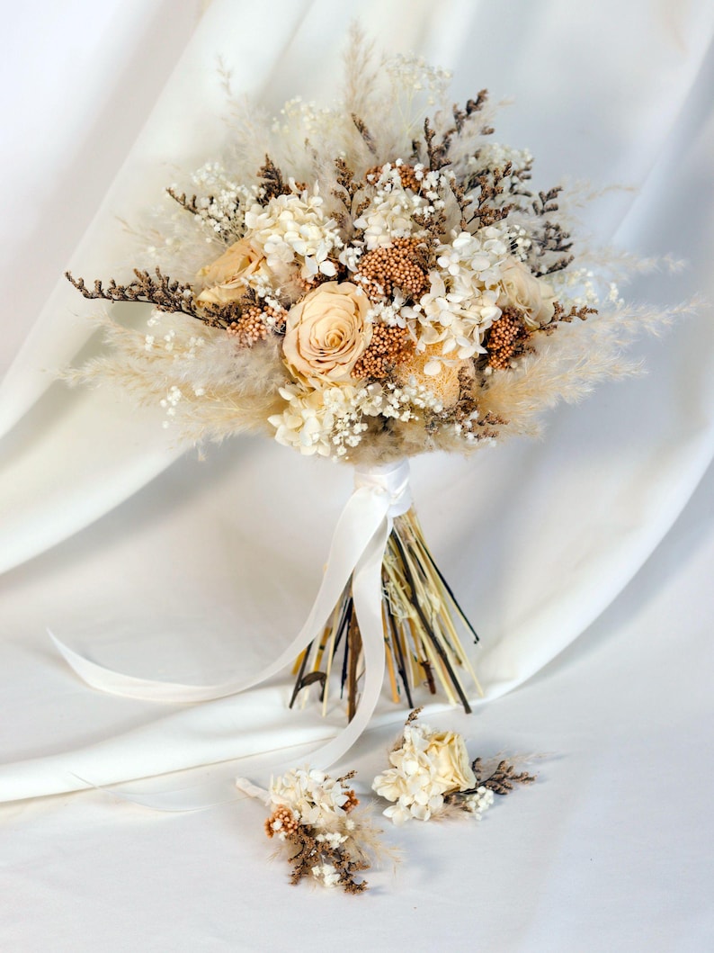 K&ouml;nnte beinhalten: Ein Brautstrau&szlig; und zwei Knopflochblumen mit Trockenblumen in Creme-, Beige- und Braunt&ouml;nen. Der Strau&szlig; enth&auml;lt Rosen, Hortensien und Pampasgras, gebunden mit einem wei&szlig;en Band. Die Knopflochblumen haben &auml;hnliche Blumenarrangements.
