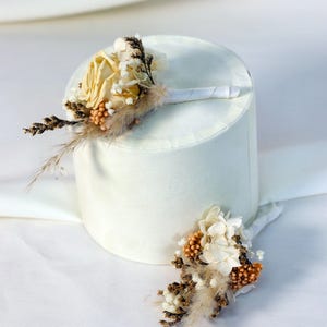 K&ouml;nnte beinhalten: Zwei Ansteckblumen mit Trockenblumen und Federn. Eine Ansteckblume hat eine blassgelbe Rose, die andere wei&szlig;e und orangefarbene Akzente. Beide sind mit wei&szlig;em Band umwickelt, ideal f&uuml;r eine Hochzeit.