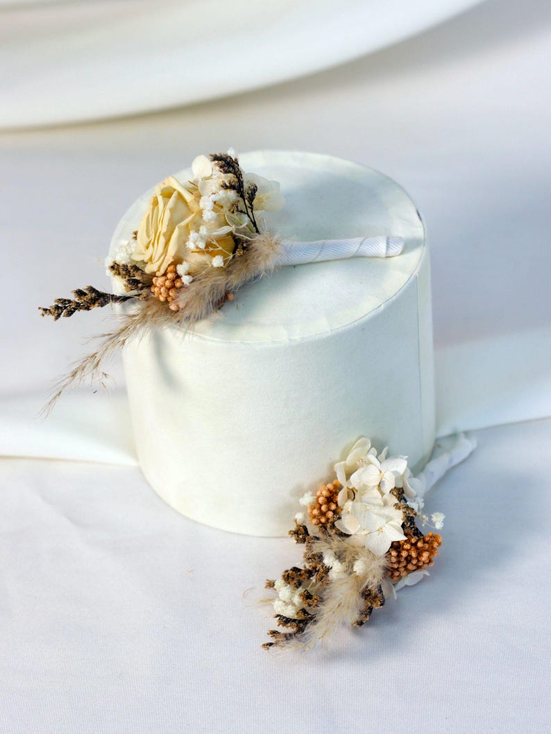 K&ouml;nnte beinhalten: Zwei Ansteckblumen mit Trockenblumen und Federn. Eine Ansteckblume hat eine blassgelbe Rose, die andere wei&szlig;e und orangefarbene Akzente. Beide sind mit wei&szlig;em Band umwickelt, ideal f&uuml;r eine Hochzeit.