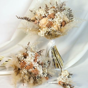 K&ouml;nnte beinhalten: Eine Sammlung von Trockenblumenstr&auml;u&szlig;en und ein Knopflochblume. Die Str&auml;u&szlig;e zeigen cremefarbene Rosen, wei&szlig;e Hortensien und flauschige beige Gr&auml;ser. Die Stiele sind mit einem wei&szlig;en Band umwickelt. Die Knopflochblume hat &auml;hnliche Blumen.