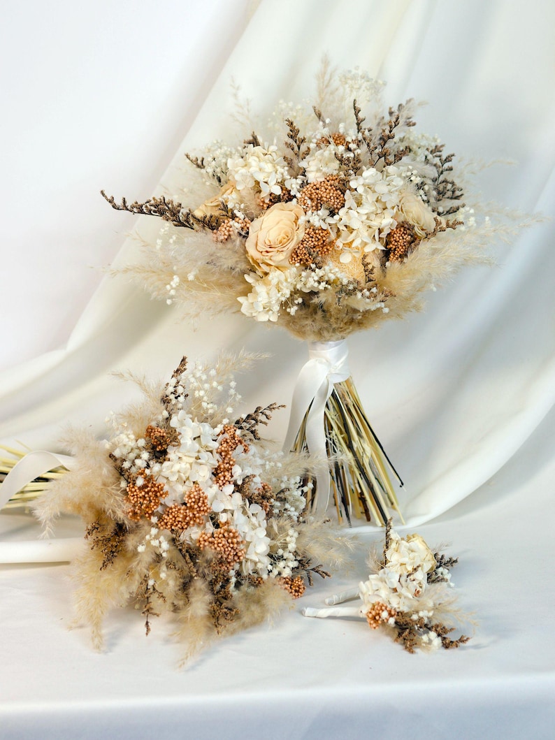 K&ouml;nnte beinhalten: Eine Sammlung von Trockenblumenstr&auml;u&szlig;en und ein Knopflochblume. Die Str&auml;u&szlig;e zeigen cremefarbene Rosen, wei&szlig;e Hortensien und flauschige beige Gr&auml;ser. Die Stiele sind mit einem wei&szlig;en Band umwickelt. Die Knopflochblume hat &auml;hnliche Blumen.