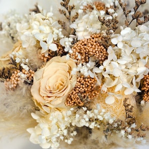 K&ouml;nnte beinhalten: Nahaufnahme eines Trockenblumenstrau&szlig;es mit einer pfirsichfarbenen Rose als Mittelpunkt. Das Arrangement umfasst wei&szlig;e Hortensien, kleine wei&szlig;e Bl&uuml;ten und braune Akzente. Der Strau&szlig; ist mit einem wei&szlig;en Band gebunden.