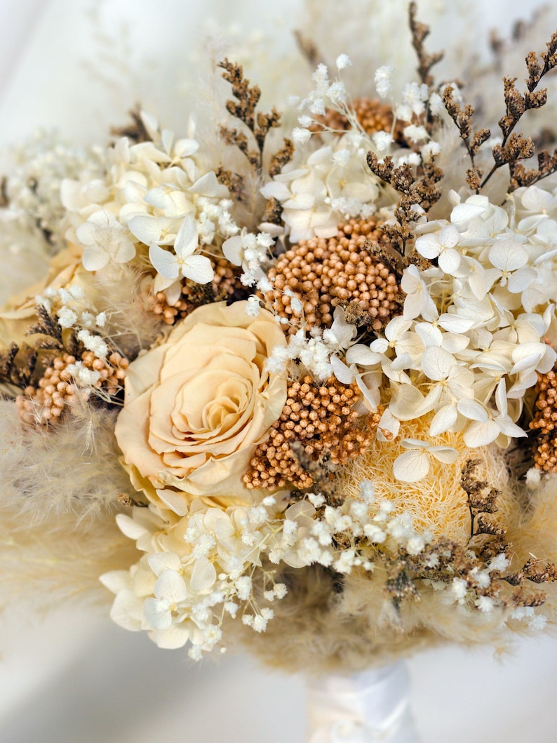 K&ouml;nnte beinhalten: Nahaufnahme eines Trockenblumenstrau&szlig;es mit einer pfirsichfarbenen Rose als Mittelpunkt. Das Arrangement umfasst wei&szlig;e Hortensien, kleine wei&szlig;e Bl&uuml;ten und braune Akzente. Der Strau&szlig; ist mit einem wei&szlig;en Band gebunden.