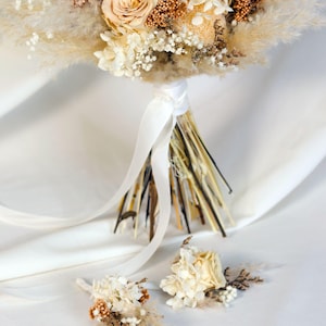 K&ouml;nnte beinhalten: Ein Brautstrau&szlig; und zwei Knopflochblumen mit Trockenblumen in Creme-, Beige- und Braunt&ouml;nen. Der Strau&szlig; enth&auml;lt Rosen, Pampasgras und andere strukturierte Elemente, gebunden mit einem wei&szlig;en Satinband. Die Knopflochblumen haben &auml;hnliche Blumengestecke.