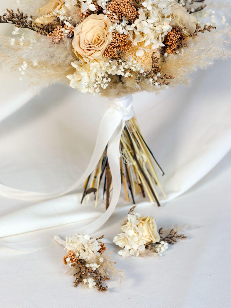 K&ouml;nnte beinhalten: Ein Brautstrau&szlig; und zwei Knopflochblumen mit Trockenblumen in Creme-, Beige- und Braunt&ouml;nen. Der Strau&szlig; enth&auml;lt Rosen, Pampasgras und andere strukturierte Elemente, gebunden mit einem wei&szlig;en Satinband. Die Knopflochblumen haben &auml;hnliche Blumengestecke.