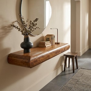 May include: A floating wooden console table with rounded edges, featuring a dark matte vase with eucalyptus branches, a stack of four hardcover books, and a slim metal desk lamp. A small wooden stool sits on the floor beneath the console.