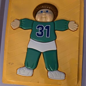 May include: A vintage Cabbage Patch Kids figure in a yellow plastic tray. The figure is wearing a green jersey with the number 31 in blue, white shorts, and white shoes. The figure has brown hair and blue eyes.