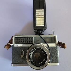 May include: Vintage Ansco camera with a black body, silver lens, and attached flash. The lens is marked "Rokkor" and "45mm f/2.8". A brown leather strap is attached to the camera body. The flash is black and rectangular.