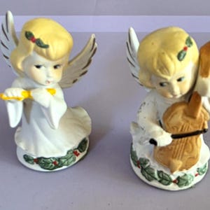 May include: Two ceramic angel figurines. One angel plays a yellow flute, the other plays a brown violin. Both angels have blonde hair, white robes, and wings. Each figurine is decorated with a holly and berry design around the base.
